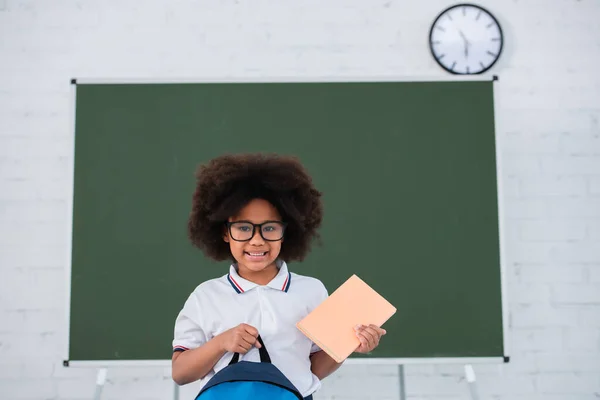 Smiling african american pupil in eyeglasses holding book and backpack near chalkboard in classroom — Stock Photo