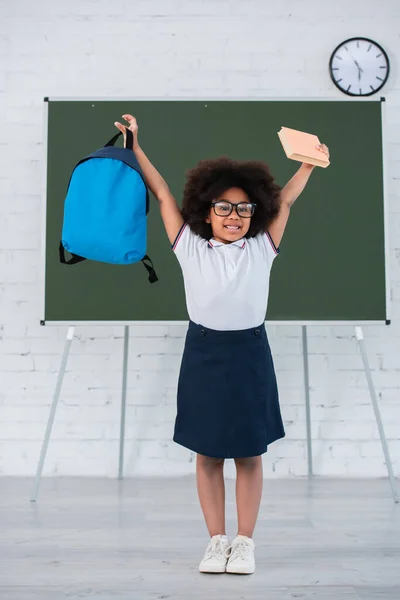 Excited african american schoolchild holding book and backpack in school — Stock Photo