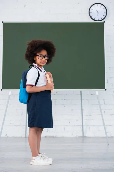 Sonriente pupila afroamericana en gafas con libro en el aula - foto de stock