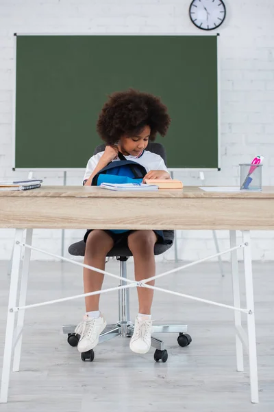 Estudante afro-americana com mochila olhando para o livro na mesa em sala de aula — Fotografia de Stock