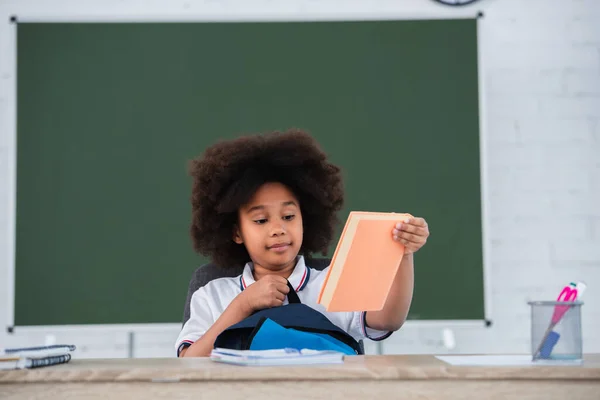 Afrikanisches Kind hält Buch und Rucksack neben verschwommenem Schreibtisch im Klassenzimmer — Stockfoto