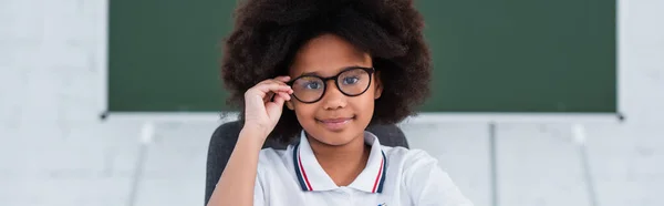 Smiling african american schoolgirl in eyeglasses looking at camera in school, banner — Stock Photo