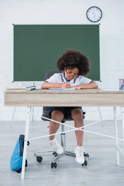 Afrikanischer Schüler mit Brille schreibt auf Notizbuch neben Rucksack im Klassenzimmer — Stockfoto