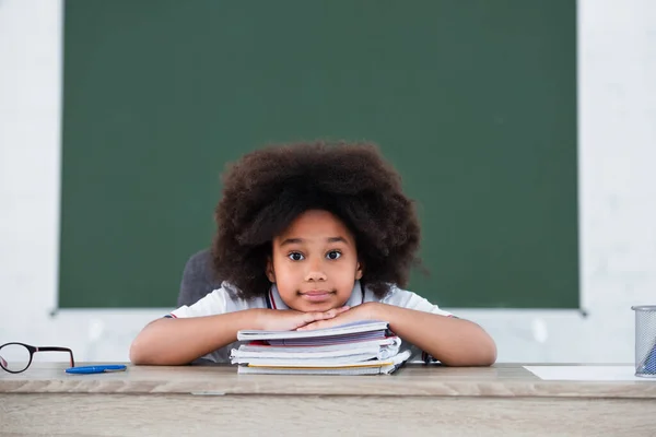 Afroamerikanisches Schulkind blickt in die Kamera in der Nähe von Notizbüchern auf dem Schreibtisch — Stockfoto