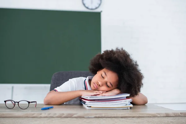 Un écolier afro-américain dormant sur des cahiers près de lunettes en classe — Photo de stock