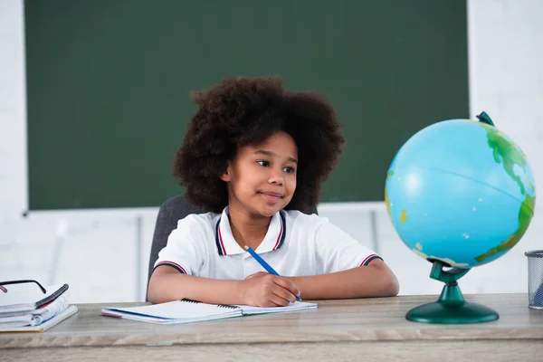 Smiling african american pupil writing on notebook near blurred globe on desk — Stock Photo