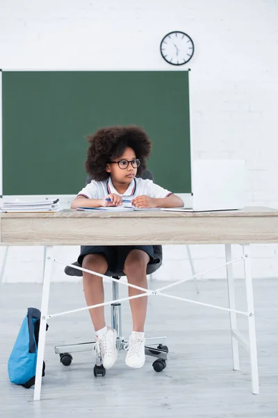 African american schoolgirl in eyeglasses holding pen near laptop and notebooks in school — Stock Photo