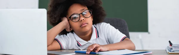 Escolar afroamericano agotado en anteojos mirando la cámara cerca de la computadora portátil y papelería, pancarta - foto de stock