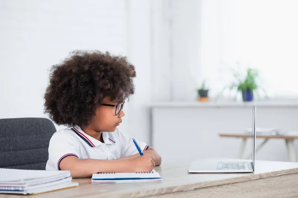 Side view of african american schoolgirl with pen and notebook near laptop in classroom — Stock Photo