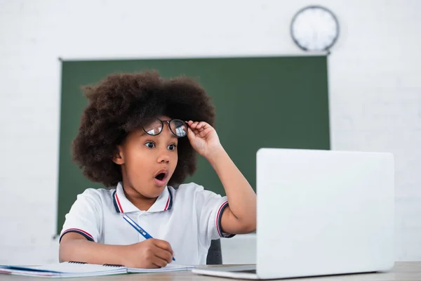 Impresionante niño afroamericano sosteniendo anteojos cerca de computadora portátil borrosa y papelería - foto de stock