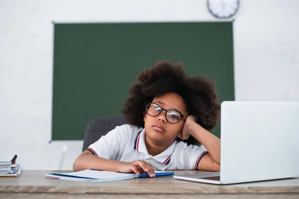 Tired african american pupil looking at camera near laptop and supplies in classroom — Stock Photo