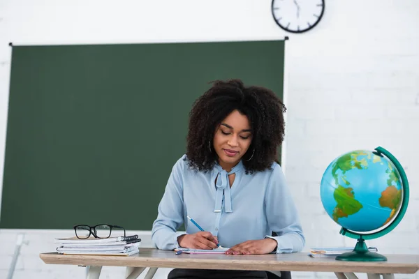 Profesora afroamericana escribiendo en cuaderno cerca del mundo en el aula - foto de stock