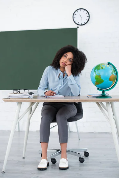 Pensive african american teacher holding pen near globe and stationery in classroom — Stock Photo