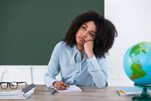 Pensive african american teacher sitting near stationery, eyeglasses and globe in classroom — Stock Photo