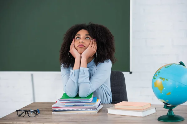 Dreamy african american teacher looking up near books, globe and copy books in classroom — Stock Photo