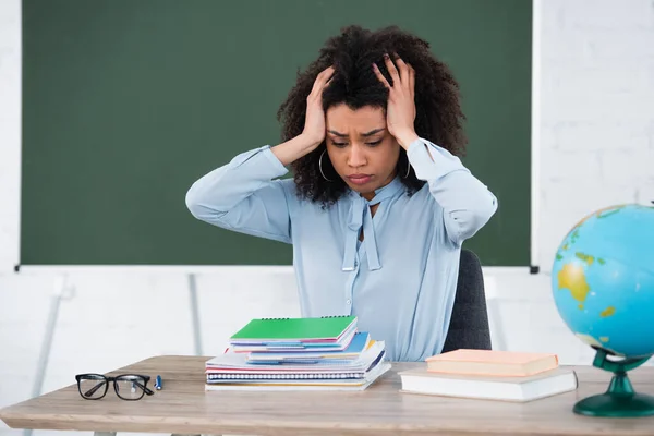 Cansado de professor afro-americano tendo dor de cabeça perto de papelaria e globo em sala de aula — Fotografia de Stock