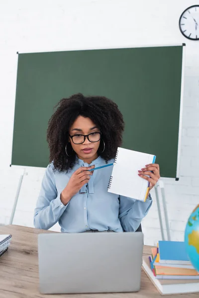 Afrikanisch-amerikanischer Lehrer zeigt bei Videoanruf im Klassenzimmer auf Notizbuch — Stockfoto