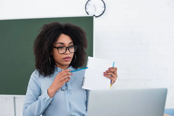 Profesora afroamericana señalando el cuaderno durante el video chat en el portátil en el aula - foto de stock