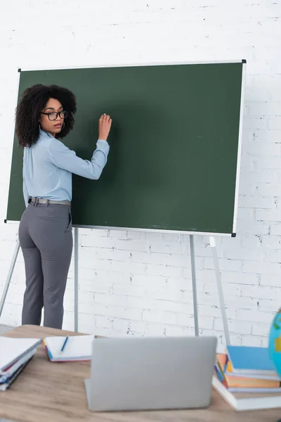 African american teacher writing on chalkboard near blurred laptop during video call in classroom — Stock Photo