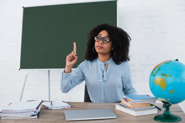 African american teacher having idea near laptop, blurred globe and stationery in classroom — Stock Photo