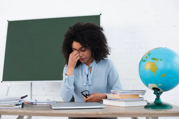 Tired african american teacher holding eyeglasses near laptop and globe in classroom — Stock Photo