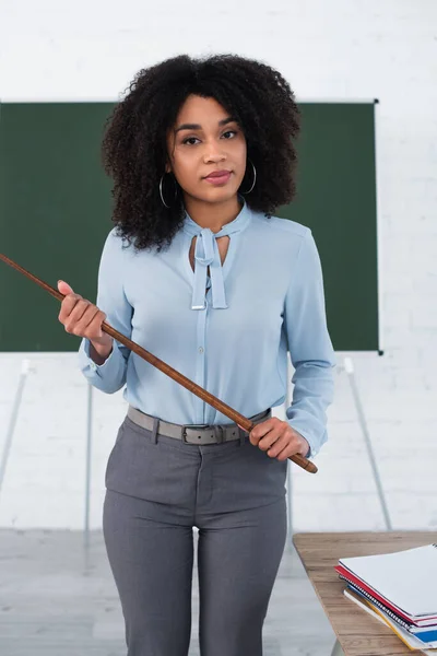 African american teacher holding pointer near notebooks in classroom — Stock Photo