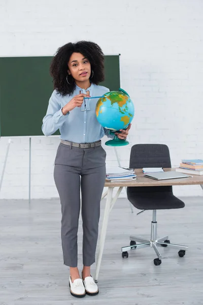Professor afro-americano apontando para o globo e olhando para a câmera na escola — Fotografia de Stock