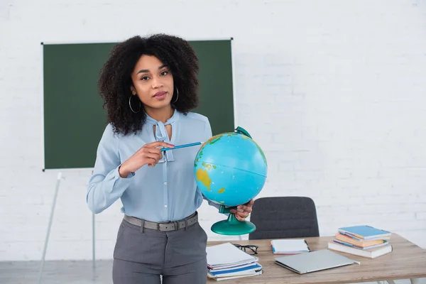 Professor afro-americano apontando para o globo e olhando para a câmera em sala de aula — Fotografia de Stock