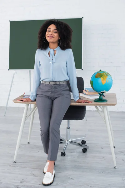 Sorrindo professor afro-americano de pé perto da mesa de trabalho em sala de aula — Fotografia de Stock