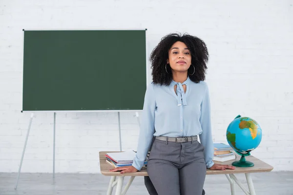 Junge afrikanisch-amerikanische Lehrerin steht neben Globus und Notizbüchern auf Arbeitstisch — Stockfoto