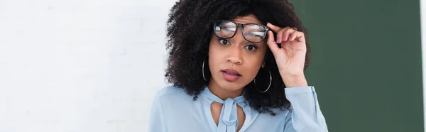 Amazed african american teacher holding eyeglasses in classroom, banner — Stock Photo