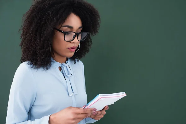 African american teacher in eyeglasses holding notebook near chalkboard — Stock Photo