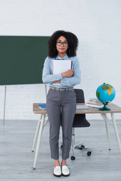 Insegnante afroamericano in occhiali che tiene quaderni vicino laptop e globo a scuola — Foto stock