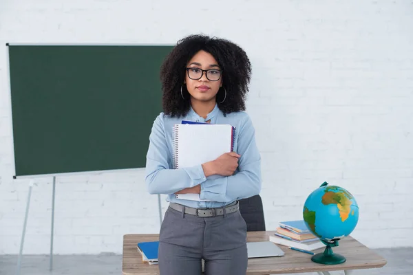 African american teacher with stationery looking at camera in classroom — Stock Photo