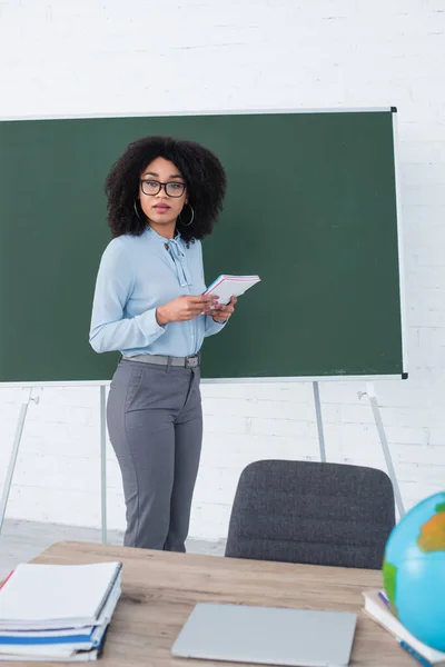 African american teacher holding notebook near chalkboard in classroom — Stock Photo