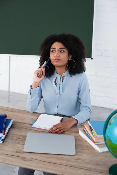 Pensive african american teacher holding pencil near notebooks and laptop on working table — Stock Photo