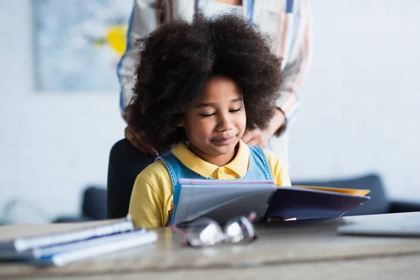 Niño afroamericano mirando el cuaderno cerca de la madre borrosa en casa - foto de stock