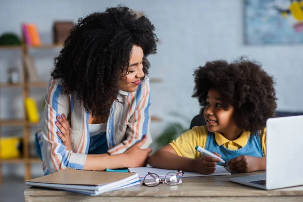 Afrikanische Mutter schaut fröhliches Kind mit Filzstift in der Nähe von Laptop und Kopierbuch an — Stockfoto