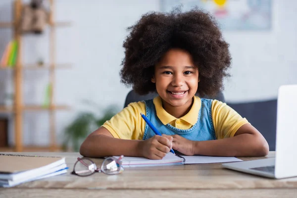 Chica afroamericana feliz escribiendo en un cuaderno cerca de la computadora portátil y anteojos en casa - foto de stock