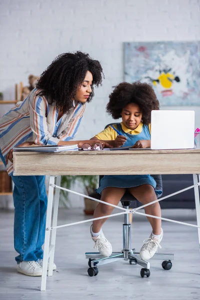 Madre afroamericana señalando el portátil cerca de la computadora portátil y la hija en casa - foto de stock