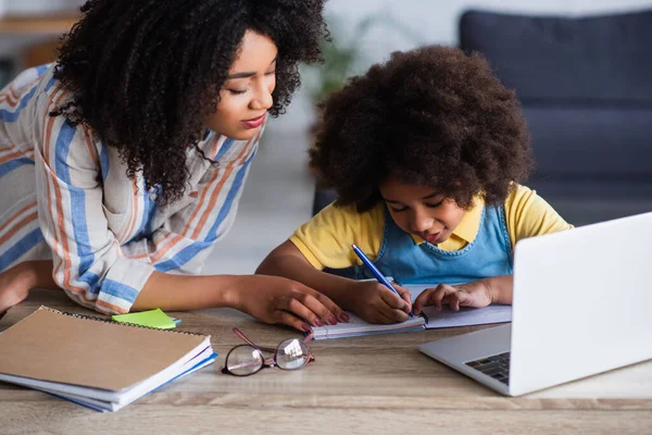 Mãe afro-americana em pé perto de criança escrevendo no notebook e laptop durante o ensino em casa — Fotografia de Stock