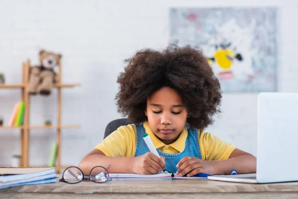 African american child with marker writing on notebook near eyeglasses and blurred laptop — Stock Photo