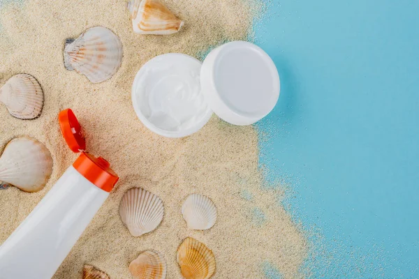 Vista dall'alto della crema cosmetica e della crema solare vicino alle conchiglie sulla sabbia e sulla superficie blu — Foto stock