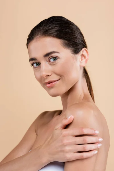 Smiling woman touching naked shoulder while looking at camera isolated on beige, beauty concept — Stock Photo