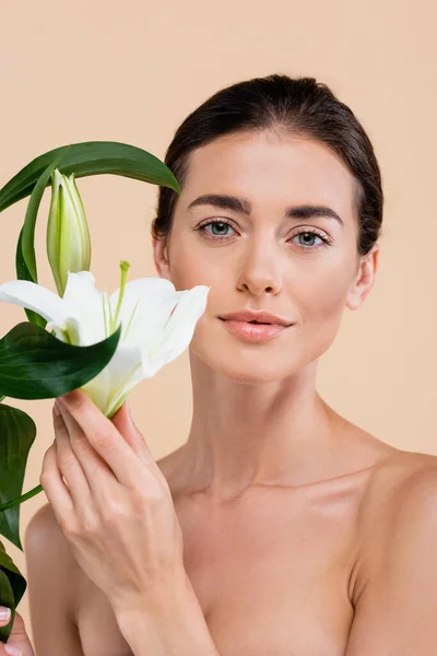 Pretty woman with naked shoulders looking at camera near lily flowers isolated on beige, beauty concept — Stock Photo