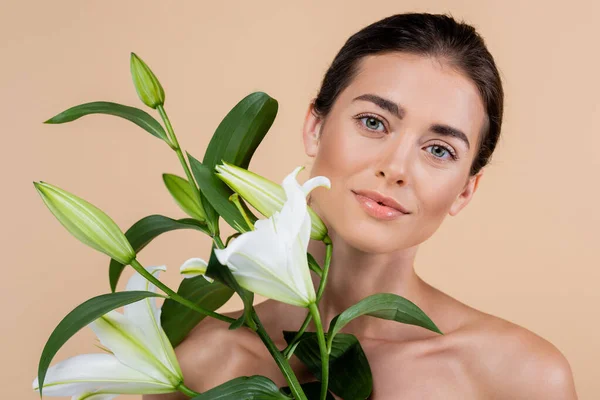 Mujer joven con la piel limpia y hombros desnudos cerca de flores de lirio aislados en beige, concepto de belleza — Stock Photo