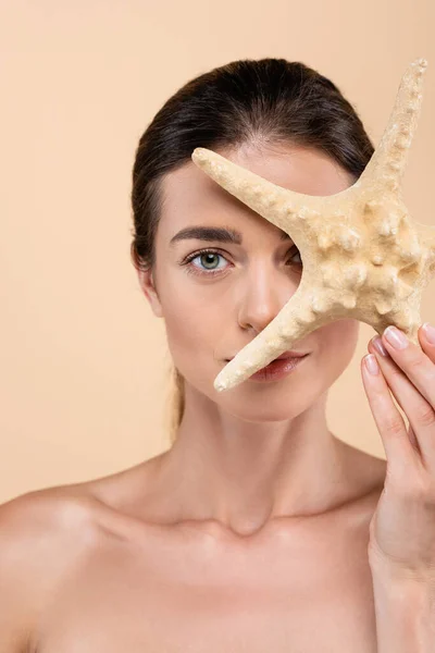 Encantadora mujer con hombros desnudos oscureciendo la cara con estrellas de mar aisladas en beige, concepto de belleza - foto de stock