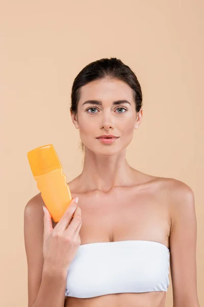 Donna bruna con spalle nude in possesso di bottiglia di crema solare isolata su beige — Foto stock