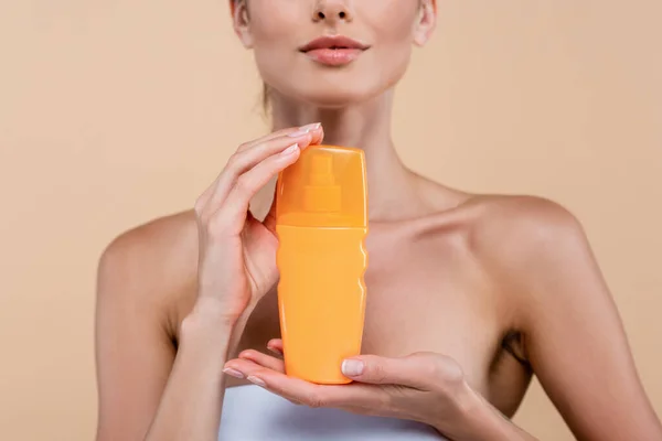 Частичный вид молодой женщины с оранжевой бутылкой крема от загара, изолированной на бежевом — стоковое фото