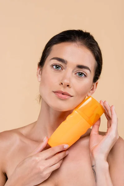 Bonita mujer joven con botella en la loción bloqueador solar mirando a la cámara aislada en beige - foto de stock